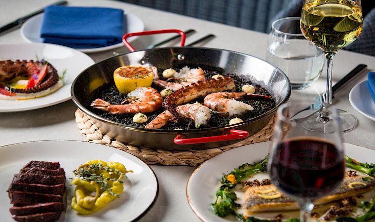 A sizzling pan of seafood and steak with lemons, served on a table with wine glasses.