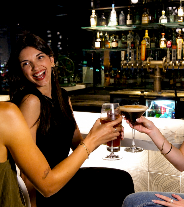 Three friends clink glasses and smile at a bar, celebrating with drinks under warm lights.