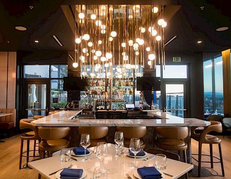 A modern restaurant/bar interior with glowing pendant globe lights, a back bar stocked with bottles, and dining tables set for guests.